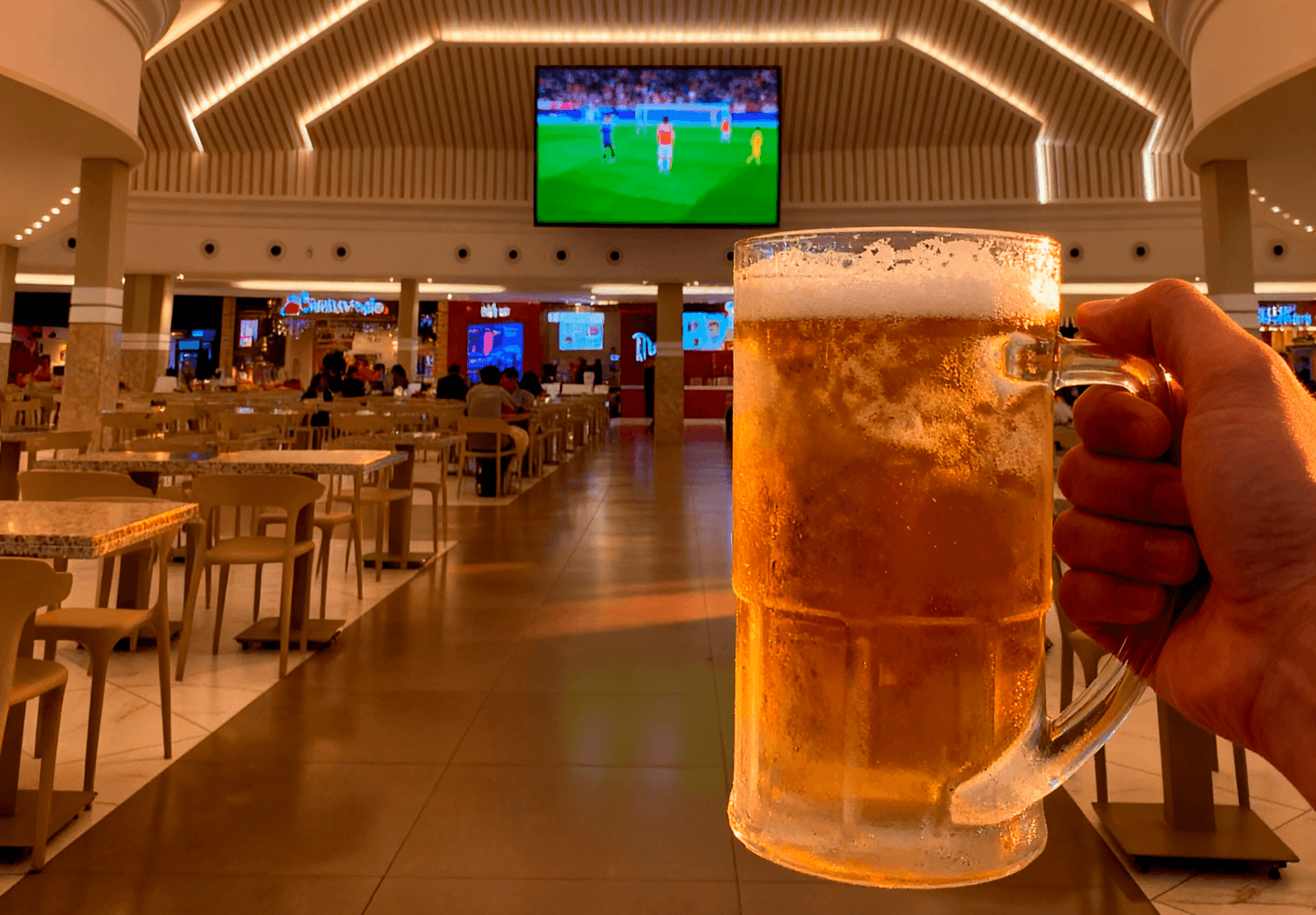 Telão exibindo jogo de futebol na praça de alimentação do Shopping Dubai, com um chopp em primeiro plano